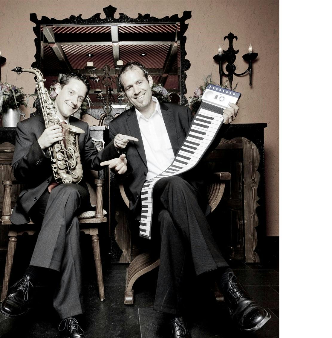 Zwei Musiker lache​ln und posieren mit einem Saxophon und einer Melodica, sitzend vor einem historischen Orgelhintergrund.