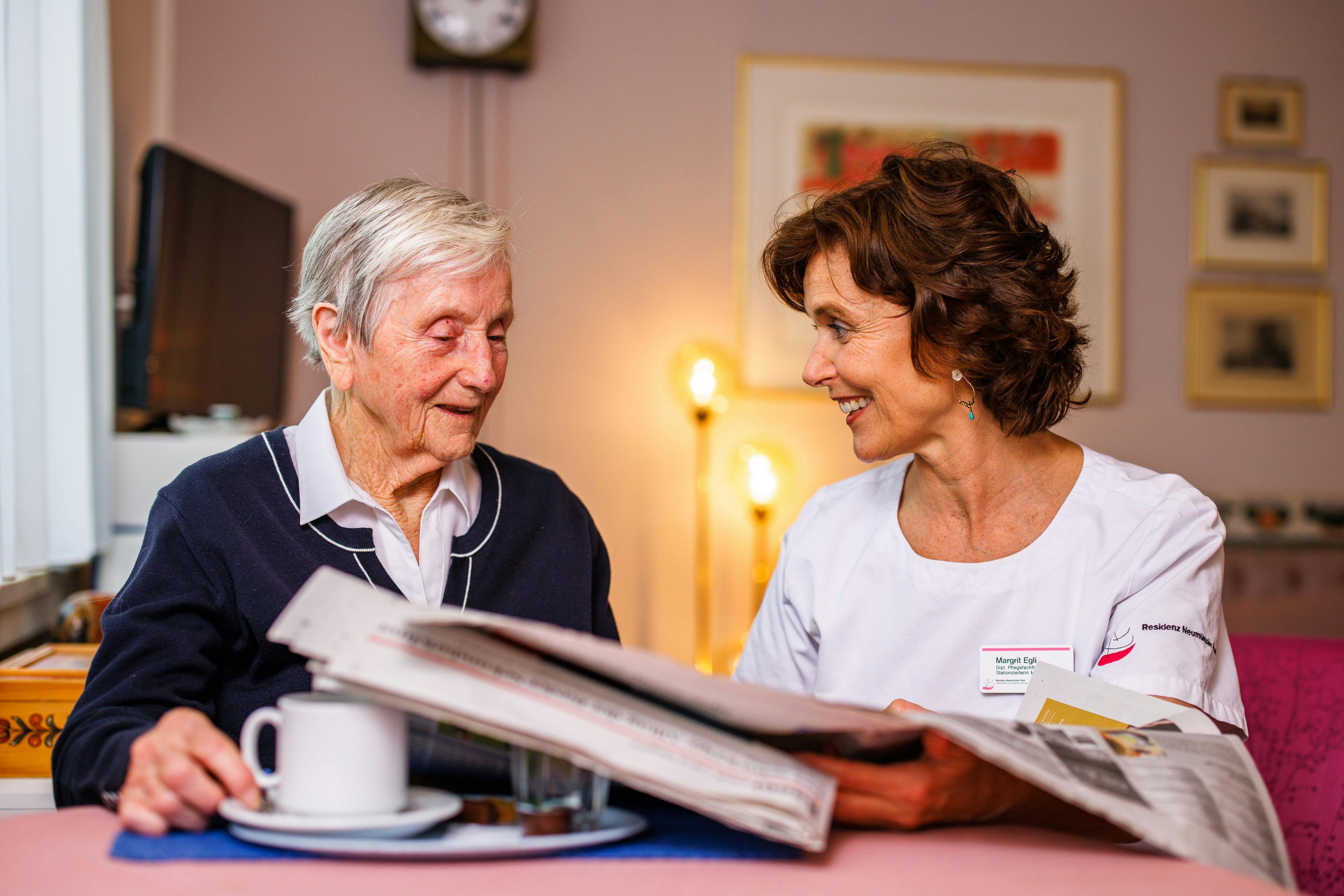 Zwei Frauen, eine ältere und eine im Pflegeberuf, lächeln sich an, während sie gemeinsam eine Zeitung betrachten.